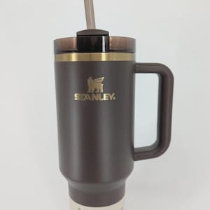 Peut inclure: Un gobelet Stanley Quencher H2.0 marron fonc&eacute; d'une capacit&eacute; de 1,18 L. Il pr&eacute;sente un logo dor&eacute;, une poign&eacute;e marron assortie, un couvercle translucide fonc&eacute; et une paille transparente. Une &eacute;tiquette &agrave; la base indique The Flowstate Quencher H2.0.