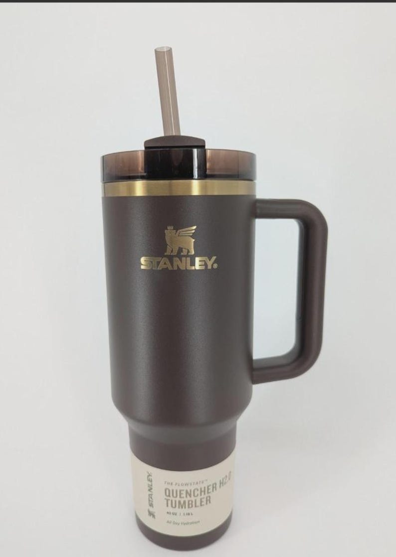 Peut inclure: Un gobelet Stanley Quencher H2.0 marron fonc&eacute; d'une capacit&eacute; de 1,18 L. Il pr&eacute;sente un logo dor&eacute;, une poign&eacute;e marron assortie, un couvercle translucide fonc&eacute; et une paille transparente. Une &eacute;tiquette &agrave; la base indique The Flowstate Quencher H2.0.