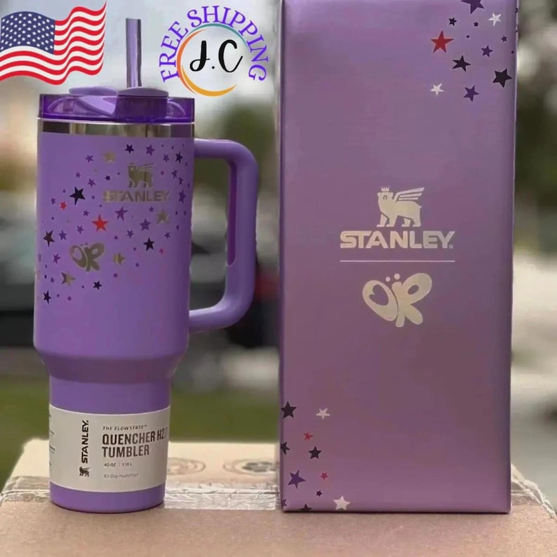 May include: A lavender Stanley Quencher H2.0 FlowState tumbler with a matching handle, straw, and lid. The tumbler features a pattern of small stars in red, purple, and silver, along with a logo. It is displayed next to its matching purple box.