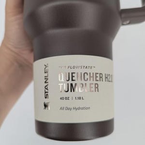 Peut inclure: Un gobelet Stanley Flowstate Quencher H2.0 marron fonc&eacute; avec une poign&eacute;e. L'&eacute;tiquette indique : Stanley, The Flowstate, Quencher H2.0 Tumbler, 40 oz | 1.18 L, All Day Hydration. Le gobelet pr&eacute;sente une finition mate et une poign&eacute;e lat&eacute;rale robuste.