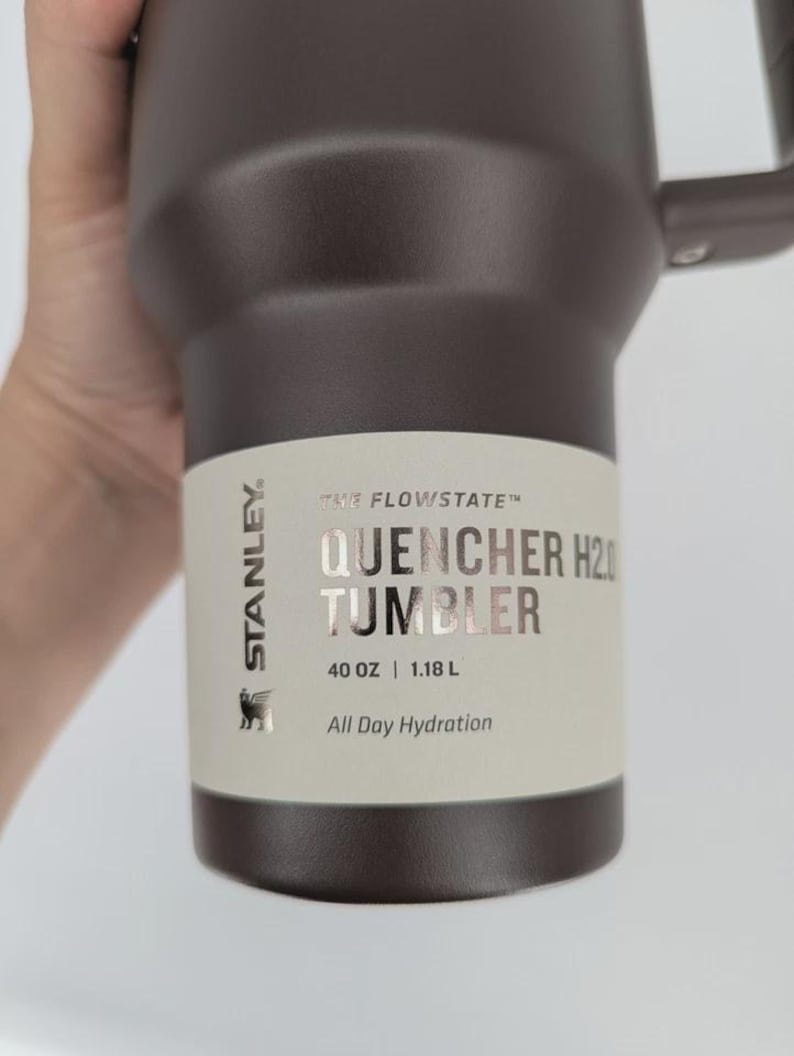 Peut inclure: Un gobelet Stanley Flowstate Quencher H2.0 marron fonc&eacute; avec une poign&eacute;e. L'&eacute;tiquette indique : Stanley, The Flowstate, Quencher H2.0 Tumbler, 40 oz | 1.18 L, All Day Hydration. Le gobelet pr&eacute;sente une finition mate et une poign&eacute;e lat&eacute;rale robuste.