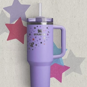 May include: A light purple stainless steel tumbler with a handle, a clear lid, and a straw. The exterior features the Stanley logo and a pattern of small stars in black, red, and purple, along with the letters OP printed on the side of the cup.