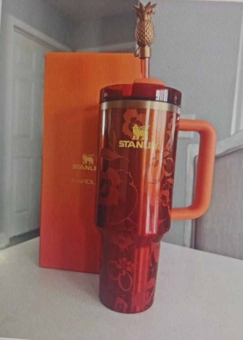 May include: A red Stanley tumbler with a floral pattern, a matching orange handle, and a lid featuring a gold pineapple straw topper. The tumbler displays the Stanley logo in gold, and an orange box with the Stanley logo and the name KAROL is behind it.