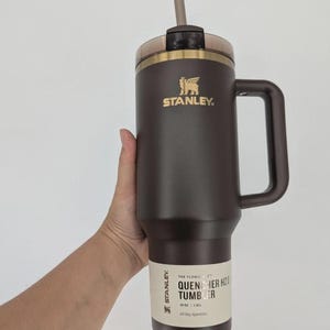 Peut inclure: Un gobelet Stanley Quencher H2.0 marron fonc&eacute; avec une poign&eacute;e, un logo dor&eacute; et une paille transparente. L'&eacute;tiquette indique : THE FLOWSTATE QUENCHER H2.0 TUMBLER 40 OZ | 1,18 L. Le gobelet poss&egrave;de un couvercle marron et un bord dor&eacute;.