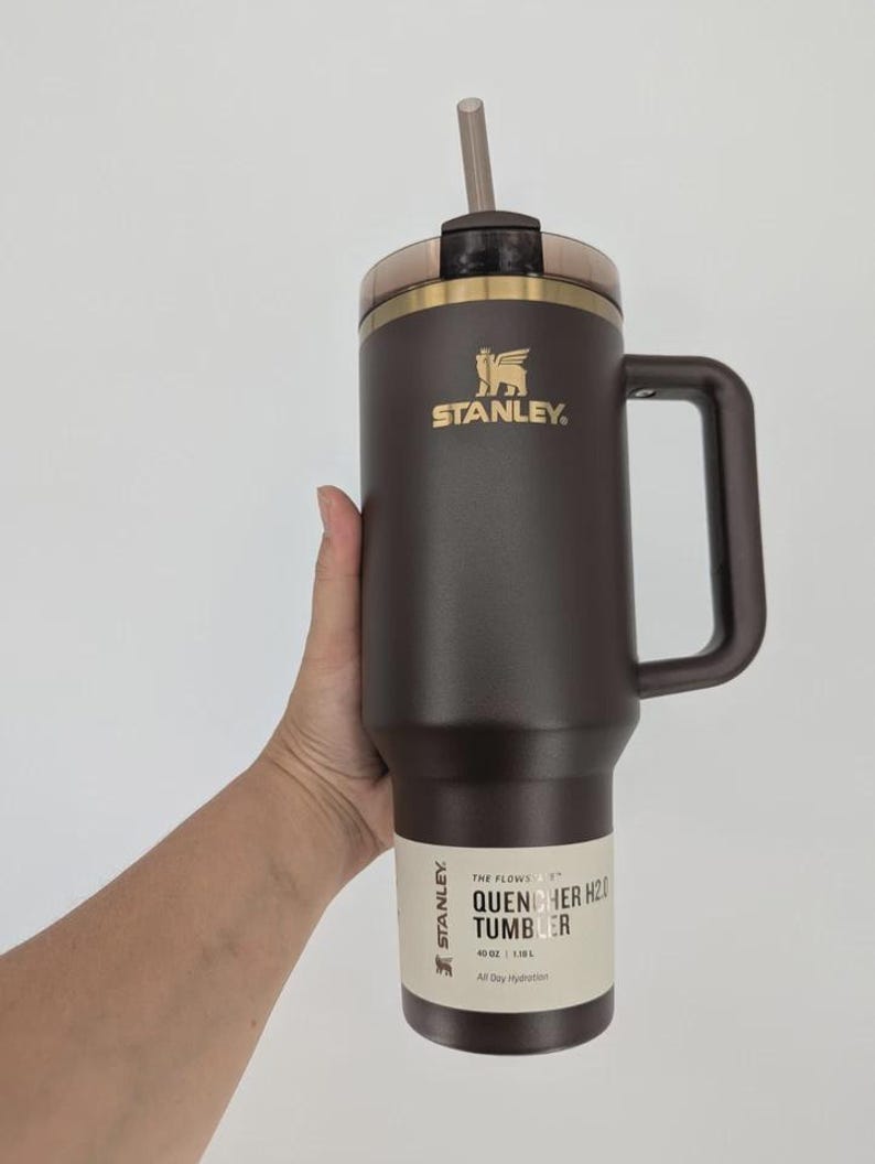 Peut inclure: Un gobelet Stanley Quencher H2.0 marron fonc&eacute; avec une poign&eacute;e, un logo dor&eacute; et une paille transparente. L'&eacute;tiquette indique : THE FLOWSTATE QUENCHER H2.0 TUMBLER 40 OZ | 1,18 L. Le gobelet poss&egrave;de un couvercle marron et un bord dor&eacute;.