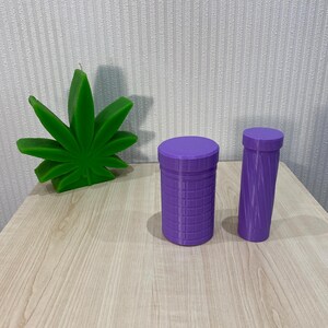 May include: A bright green candle shaped like a cannabis leaf, accompanied by two purple cylindrical containers with lids. One container features a grid-like texture on its exterior, while the other has a smooth, spiraling, twisted design.