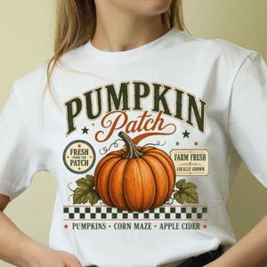 May include: A white short-sleeve t-shirt featuring a graphic of an orange pumpkin with green leaves. The text reads PUMPKIN PATCH, FRESH FROM THE PATCH, FARM FRESH LOCALLY GROWN, PUMPKINS, CORN MAZE, and APPLE CIDER above a black and white checkered border.