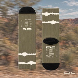 Könnte beinhalten: Ein Paar olivgrüne Crew-Socken mit schwarzen Bündchen und Fersen sowie weißen horizontalen Tie-Dye-Streifen. Auf dem Stoff befindet sich weißer Text, darunter das DC-Logo, Materialangaben und Pflegehinweise für die Socken.
