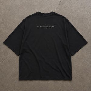 Könnte beinhalten: Ein schwarzes Kurzarm-T-Shirt mit Rundhalsausschnitt und lockerer Passform. Auf dem oberen Rücken befindet sich der weiße Schriftzug "NO GLORY IN COMFORT" in einer klaren serifenlosen Schrift. Das Kleidungsstück besteht aus einfarbigem Stoff.