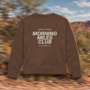May include: A brown crewneck sweatshirt featuring white text on the back that reads MADE FOR RUNNING MORNING MILES CLUB and DAILY CHORE RUNNING CLUB. The garment has long sleeves, a ribbed neckline, and a ribbed hem, presented in a solid color.