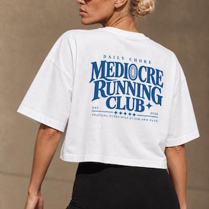 May include: A white cropped t-shirt featuring blue text on the back that reads DAILY CHORE MEDIOCRE RUNNING CLUB EST. 2024 ENJOYING EVERY MILE AT OUR OWN PACE. The shirt has short sleeves and a boxy fit, paired with solid black leggings.