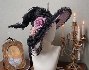May include: A black witch hat featuring a wide brim with black lace trim, adorned with large purple and light pink fabric roses, black feathers, and a dangling black beaded fringe. The underside of the brim is lined with light purple fabric.