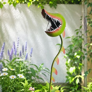 May include: A metal sculpture of a Venus flytrap plant with a green and red open mouth featuring sharp teeth. The plant has a long, slender green stem with small red-tipped leaves and is planted in a simple, neutral-colored cylindrical pot.