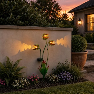 May include: A decorative garden light shaped like a three-headed Venus flytrap plant with green stems and leaves. Each head features an open mouth with jagged teeth and a warm, glowing yellow light inside, casting a distinct shadow on the wall.
