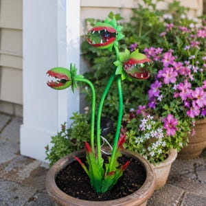 May include: A decorative garden stake featuring three green and red carnivorous plant heads with sharp teeth, mounted on thin green stems. The plant heads are positioned at varying heights, and the base includes a small black solar panel component.