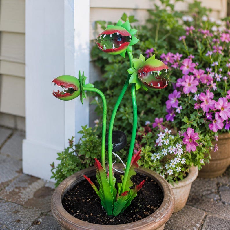 May include: A decorative garden stake featuring three green and red carnivorous plant heads with sharp teeth, mounted on thin green stems. The plant heads are positioned at varying heights, and the base includes a small black solar panel component.