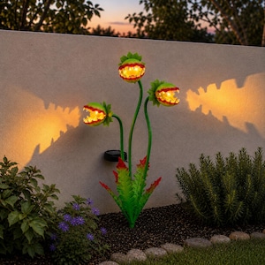 May include: A solar-powered garden light shaped like a three-headed Venus flytrap plant. The green leaves have red-tipped edges, and each open mouth features a glowing yellow light, casting a warm, bright orange shadow against the wall behind.