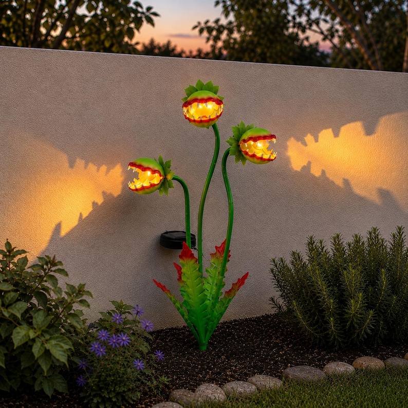 May include: A solar-powered garden light shaped like a three-headed Venus flytrap plant. The green leaves have red-tipped edges, and each open mouth features a glowing yellow light, casting a warm, bright orange shadow against the wall behind.