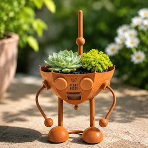 May include: A rust-colored metal planter shaped like a robot with two legs, two arms, and a central antenna. The front features the text "PLANT BUDDY" with a small leaf icon above a vent detail. It holds a green succulent and a small shrub.