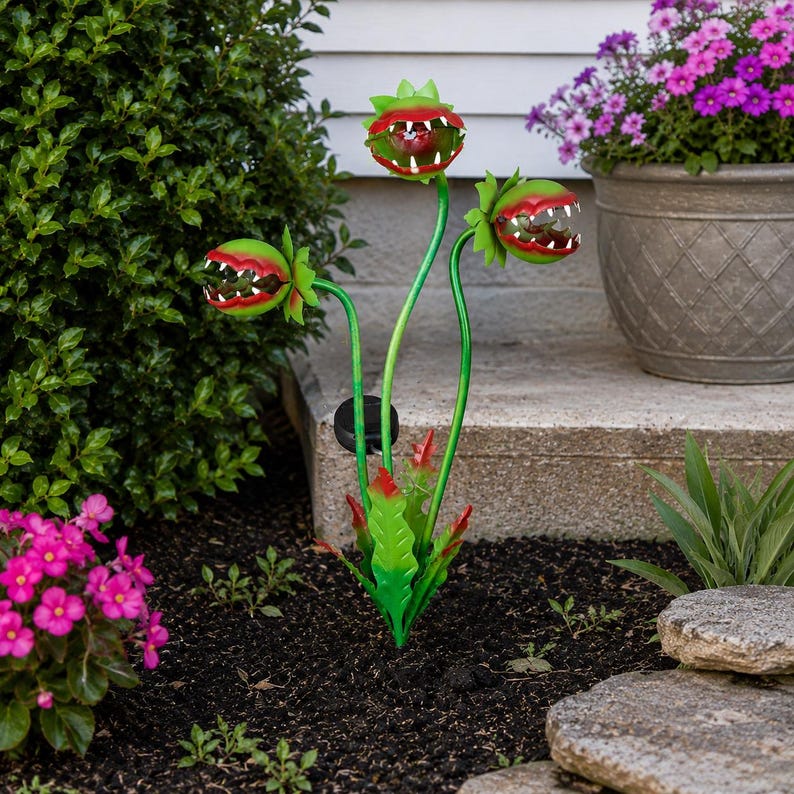 May include: A metal garden stake featuring three green Venus flytrap heads with red interiors and white teeth. The plant sculpture has long, slender green stems and jagged green leaves at the base, with a small black solar panel attached to one stem.