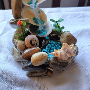 May include: A decorative miniature fountain made of seashells, stones, and small blue stones. The fountain features a cascading water effect with a small orange flower and green plants. The base is composed of various sized stones.