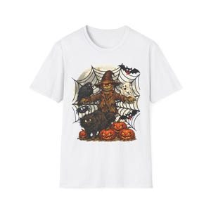 May include: A white short-sleeve t-shirt featuring a graphic print of a scarecrow, a black cat, a ghost, a crow, a spider, and three orange jack-o'-lanterns set against a large spider web background with a full moon and flying bats.