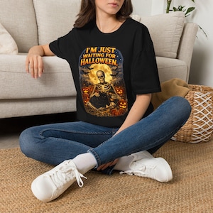 May include: A black t-shirt featuring a graphic print of a skeleton sitting in a pumpkin patch under a full moon with bats. The text "I'M JUST WAITING FOR HALLOWEEN" is printed in orange above the skeleton. The shirt has short sleeves and a crew neck.