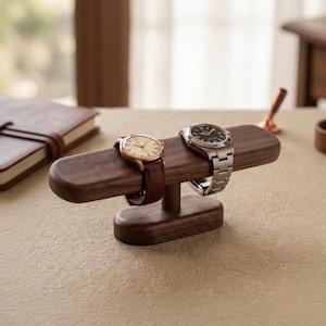 Peut inclure: Un présentoir à montres en bois brun foncé composé d'une barre horizontale soutenue par un pilier central sur une base ovale. Deux montres-bracelets sont exposées sur la barre : une avec un cadran crème et un bracelet en cuir brun, et une avec un cadran noir et un bracelet à maillons en métal argenté.