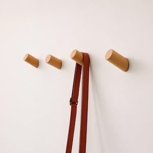 Peut inclure: Quatre patères cylindriques en bois clair fixées en ligne horizontale. Une longue sangle fine de couleur brun rougeâtre foncé est suspendue à la troisième patère en partant de la gauche. Les patères ont une finition lisse en bois naturel.