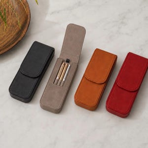 Peut inclure: Quatre étuis à stylos rectangulaires de couleur noire, taupe, fauve et rouge. L'étui taupe est ouvert, révélant trois stylos métalliques à l'intérieur. Chaque étui présente une fermeture à rabat et une finition mate lisse.