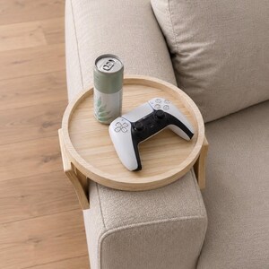 Peut inclure: Un plateau d'accoudoir rond en bois clair avec des rabats latéraux articulés conçus pour se fixer sur l'accoudoir d'un canapé. Le plateau supporte une manette de jeu sans fil blanche et noire et une canette cylindrique avec un motif de feuilles vertes.