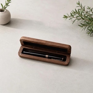 Peut inclure: Un étui à stylo rectangulaire en bois avec un couvercle à charnière, présentant une finition en grain de bois brun foncé. À l'intérieur de l'étui ouvert repose un stylo à bille noir élégant avec des accents dorés sur la bague et le clip.
