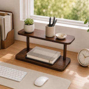 May include: A two-tiered dark wood desk organiser with rounded edges. The top shelf holds a small potted succulent, a cylindrical pen holder with three pens, and a small white dish. A grey notebook rests on the bottom shelf of the wooden stand.