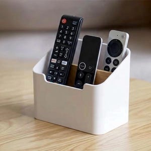 Puede incluir: Un organizador de escritorio de plástico blanco con un divisor interior de madera clara, que contiene tres mandos a distancia. El organizador presenta un borde frontal curvo y una base rectangular para guardar mandos.