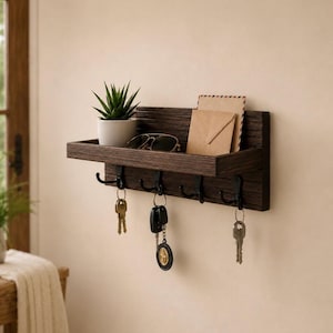 Peut inclure: Une étagère murale en bois brun foncé dotée de quatre crochets en métal noir. L'étagère supporte une petite plante succulente verte dans un pot blanc, une paire de lunettes de soleil et deux enveloppes beiges. Trois trousseaux de clés pendent.