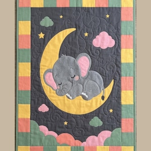Pode incluir: Uma colcha retangular com um painel central cinza-escuro apresentando um elefante cinza dormindo em uma lua crescente amarela. O design inclui nuvens rosas, estrelas amarelas e nuvens verdes. A borda consiste em quadrados alternados amarelos, verdes e rosas.