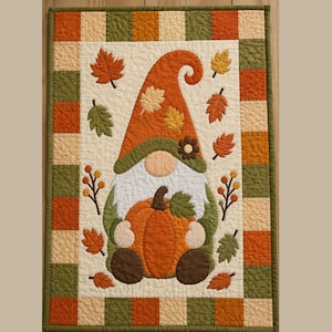 Peut inclure: Une tenture murale matelassée rectangulaire représentant un gnome central portant un grand chapeau orange et tenant une grosse citrouille orange. Le motif est entouré de feuilles d'automne éparpillées et d'une bordure à carreaux orange, vert et crème.