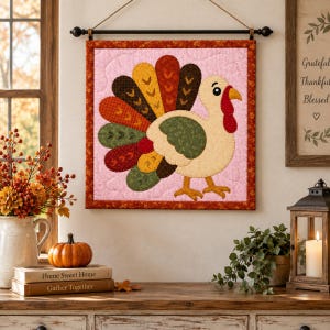 May include: A square fabric wall hanging featuring a quilted turkey with a colorful tail of patterned feathers in shades of red, orange, yellow, brown, and green. The turkey is set against a light pink background with a dark red patterned border.