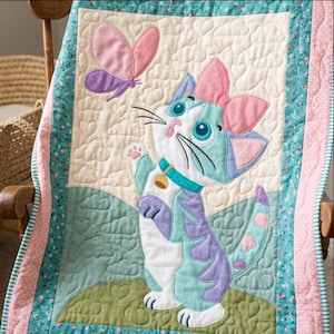 Op de afbeelding: Een gewatteerde babydeken met een centrale applicatie van een wit, groenblauw en paars kitten met een roze strik, reikend naar een roze en paarse vlinder. De achtergrond heeft een hartjes- en wolkenpatroon met bloemrijke randen in groenblauw en roze.