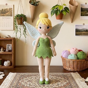 May include: A handmade crochet doll of a fairy with yellow hair, a green dress, and blue wings. The doll is standing on a patterned rug. The background includes plants, a calendar, and a basket of yarn.