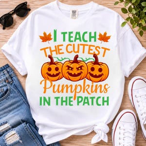 May include: A white short-sleeve t-shirt featuring a graphic print with three orange jack-o'-lantern pumpkins and the text "I TEACH THE CUTEST PUMPKINS IN THE PATCH" in orange and green lettering, accented by two small orange maple leaves.