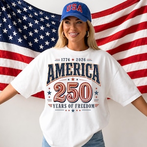 Pode incluir: Uma t-shirt branca de manga curta com um gráfico contendo o texto 1776 2026 AMERICA 250 YEARS OF FREEDOM em azul e vermelho, acompanhado de estrelas. Um boné azul com o texto vermelho USA é usado acima da t-shirt.