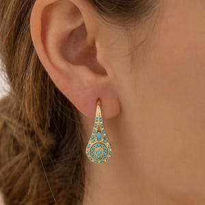 May include: A gold-toned drop earring featuring a teardrop-shaped frame adorned with small, round, bright turquoise stones. The design includes a central circular motif surrounded by intricate metalwork and a secure hook-style closure.
