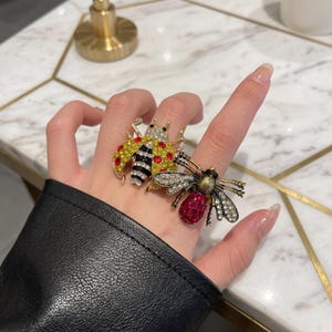 May include: Two statement rings featuring insect designs. One ring is a ladybug with yellow and red rhinestones and black stripes. The second ring is a bee with clear crystal wings and a red faceted stone body, both set in a metallic gold tone.