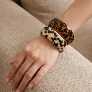 May include: Two wide bangle bracelets worn together. One bracelet features a dark brown and black tortoiseshell pattern, while the other displays a cream and black spotted animal print. Both bangles have a smooth, glossy, rounded surface.