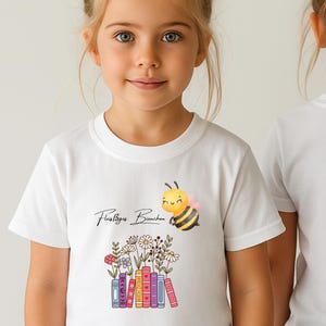 Könnte beinhalten: Ein weißes T-Shirt mit kurzen Ärmeln, das mit einer lächelnden Biene, dem Schriftzug "Fleißiges Bienchen" und einer Reihe bunter Bücher bedruckt ist, aus denen verschiedene Wildblumen wie Gänseblümchen in Rosa, Blau, Gelb und Rot wachsen.