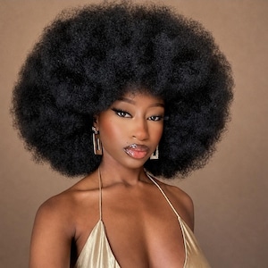 Könnte beinhalten: Eine Person mit einer voluminösen, dunklen, texturierten Afro-Frisur, die goldfarbene, rechteckige Ohrhänger und ein metallisch goldenes Neckholder-Oberteil trägt. Die Person hat dunklen Eyeliner, definierte Augenbrauen und glänzende Lippen.