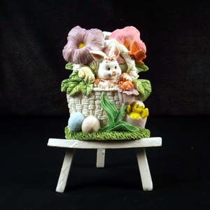 May include: A white bunny rabbit figurine sits in a woven basket with pink, purple, and yellow flowers. The basket is on a white easel.