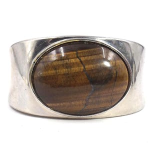 Puede incluir: Un anillo ancho de plata con una gran piedra ojo de tigre ovalada. La piedra es de color marrón cálido con estrías naturales. El anillo tiene un diseño simple y pulido, que muestra la gema.