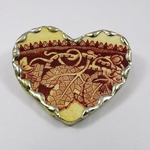Puede incluir: Un broche en forma de corazón con un patrón floral rojo y blanco, rodeado por un borde de color plateado.